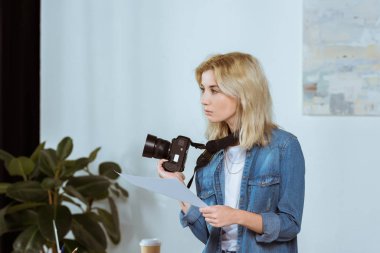 side view of thoughtful photographer with photo camera and photoshoot example looking away in studio clipart