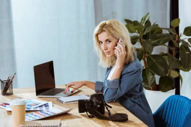 smartphone üzerinde konuşmak ve Studio dizüstü bilgisayar ile işyeri kameraya bakarak genç fotoğrafçı yan görünüm