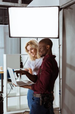 african american photographer and caucasian model choosing photos on laptop together during photoshoot in studio clipart