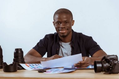 portrait of smiling african american photographer sitting at workplace with photoshoot examples and photo camera and looking at camera clipart