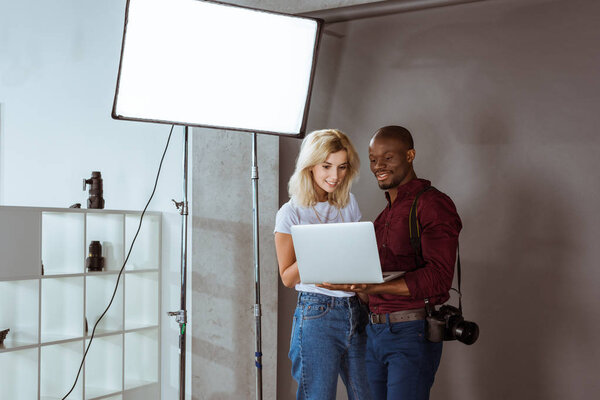 african american photographer and caucasian model choosing photos on laptop together during photoshoot in studio