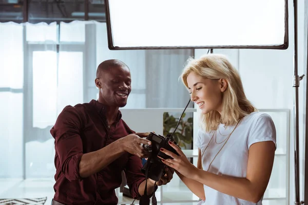 陽気なアフリカ系アメリカ人カメラマンとスタジオで撮影中に写真を一緒に選択する白人モデルの肖像 — ストック写真