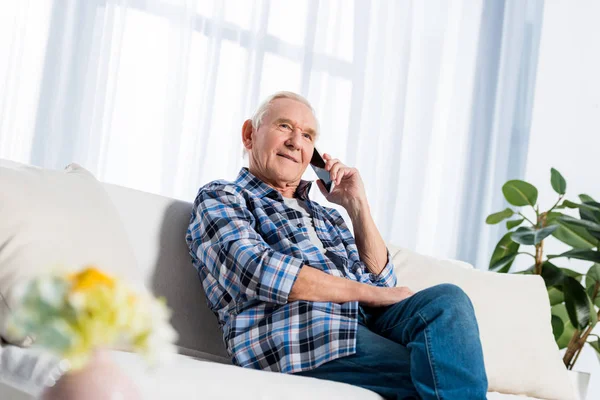 pensive senior man talking on smartphone while sitting on sofa at home