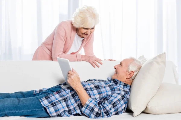 老人与片剂看微笑的妻子附近 当休息在沙发在家时 — 图库照片