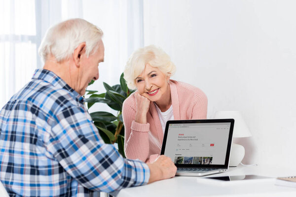 portrait of smiling senior woman looking at husband using laptop with airbnb logo at home