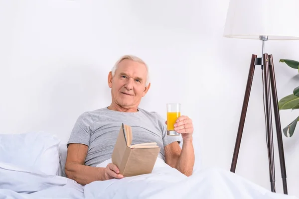 portrait of senior man with glass of juice and book in hands looking at camera while lying in bed at home