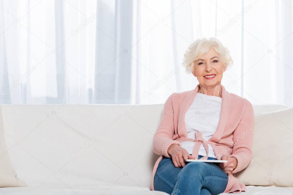 portrait of smiling senior woman with tablet in hands resting on sofa and looking at camera at home