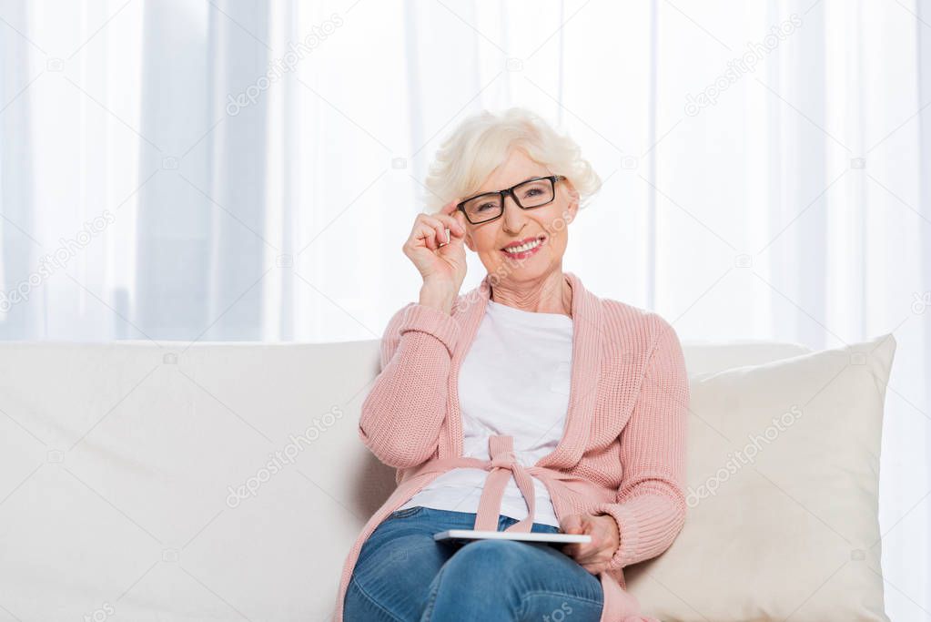 portrait of smiling senior woman in eyeglasses with tablet in hands resting on sofa and looking at camera at home