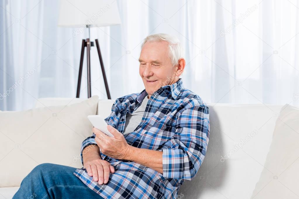 portrait of smiling senior man sitting on sofa and using smartphone at home