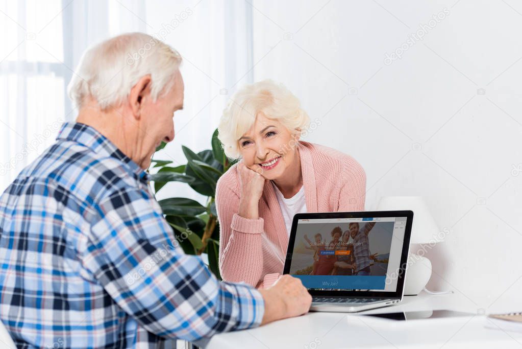 Smiling senior woman looking at husband that using laptop at home