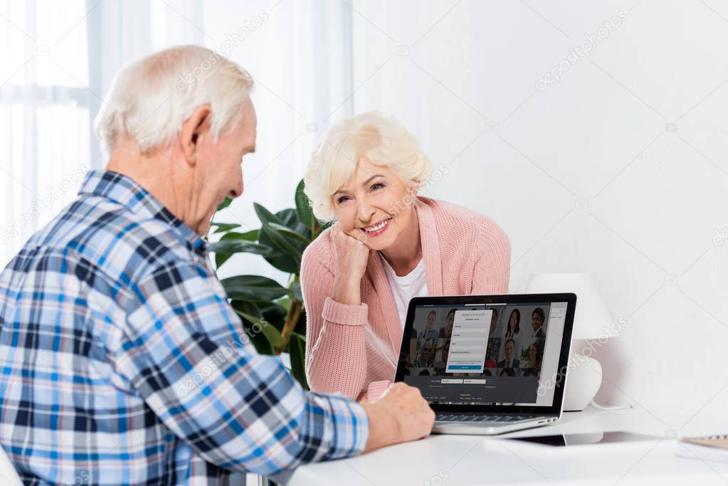 Portrait of smiling senior woman looking at husband using laptop at home