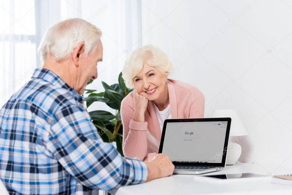 Portrait of cheerful senior woman looking at husband working on laptop at home