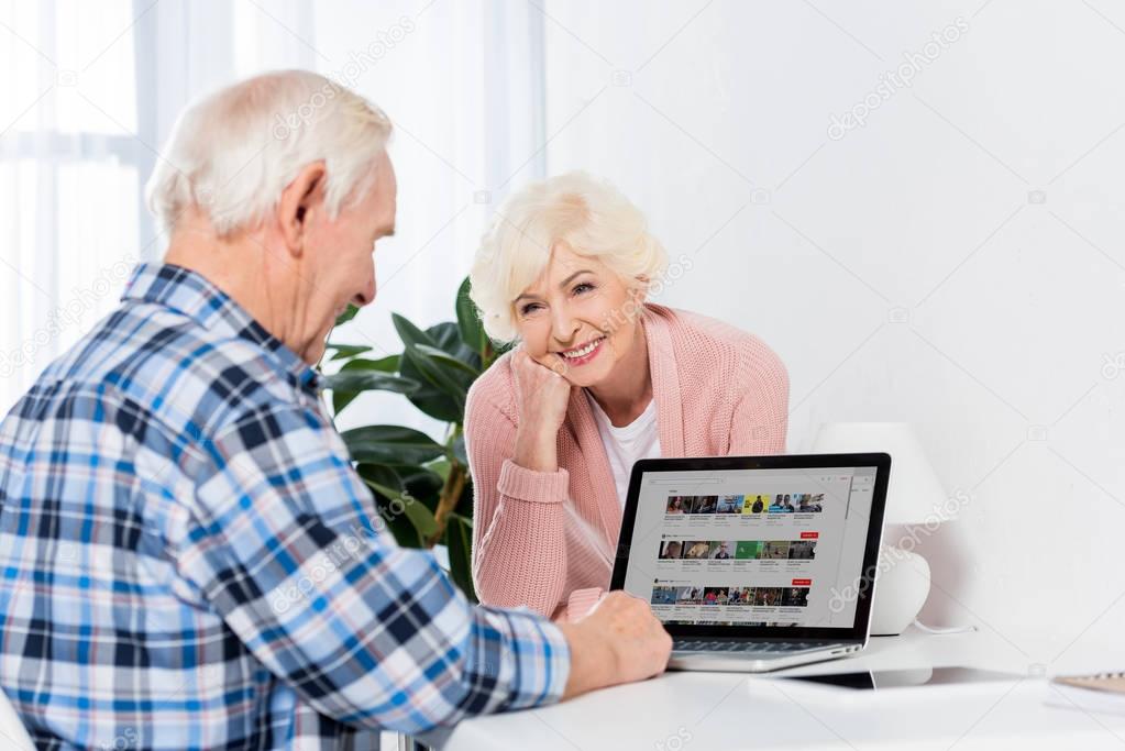 Portrait of cheerful senior woman looking at husband using laptop at home
