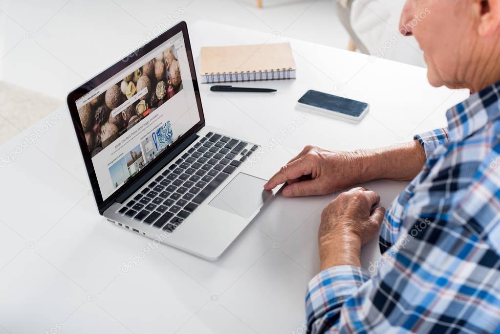 Partial view of senior man working on laptop with depositphotos logo at table with notebook at home
