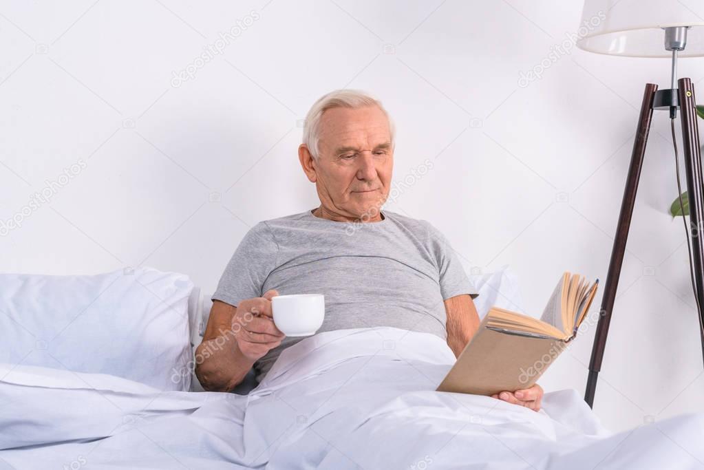 portrait of senior man with cup of coffee reading book in bed at home