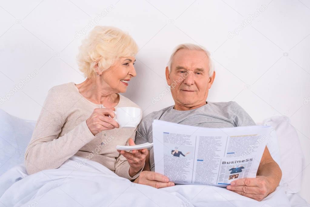 portrait of senior woman drinking coffee while husband reading newspaper in bed at home