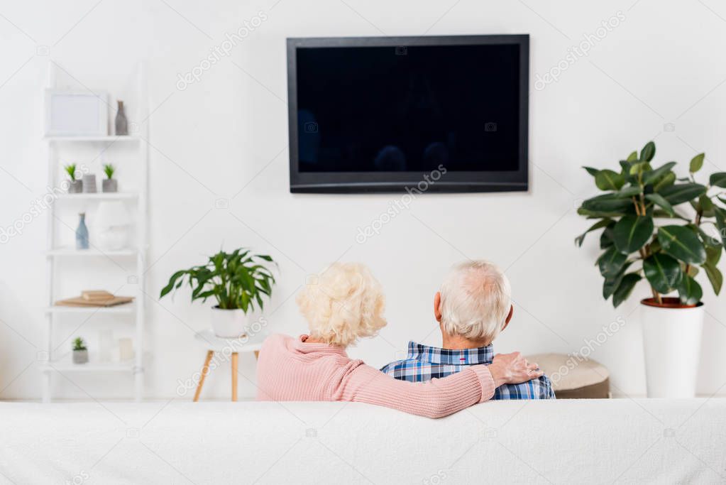 back view of senior couple watching tv and embracing on couch