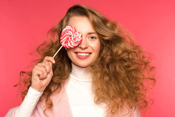 smiling girl covering eye with heart shaped lollipop isolated on red