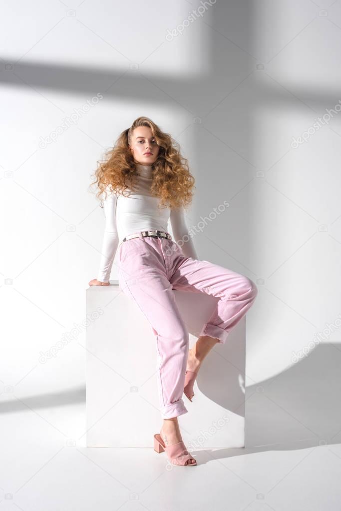 attractive serious woman with curly hair sitting on white cube and looking at camera 