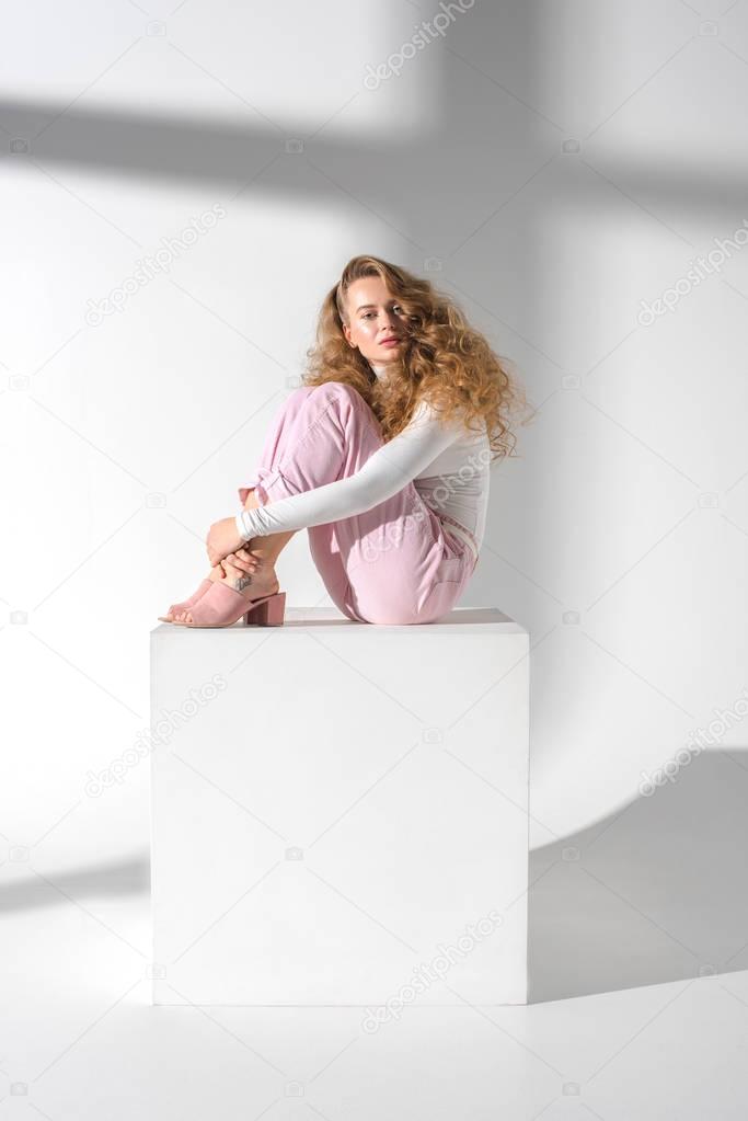 beautiful serious woman with curly hair sitting on white cube and hugging legs