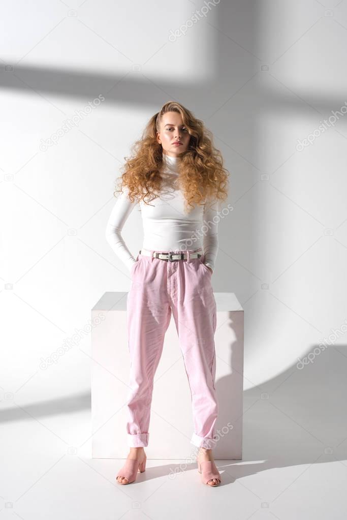 serious stylish girl with curly hair standing in front of white cube