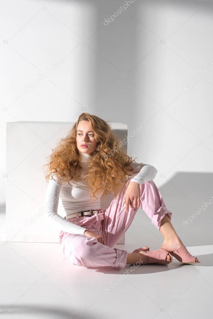 confident stylish girl sitting on a floor near white cube and looking away 