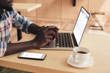 facebook messenger ve kafede Web sitesi facebook ile dizüstü bilgisayar ile akıllı telefon kullanan Afrikalı-Amerikalı adam görünümünü kırpılmış
