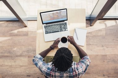 Afrika kökenli Amerikalı adam kahve içme ve dizüstü bilgisayar ile couchsurfing Web sitesi kullanarak havai görünümü