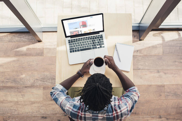 над головой вид африканского американца, пьющего кофе и пользующегося ноутбуком с веб-сайтом ebay
