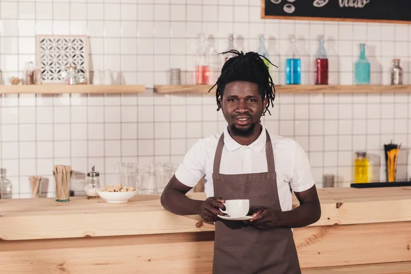 バーのカウンターに立っているコーヒー カップを保持しているエプロンのアフリカ系アメリカ人のバリスタ — ストック写真