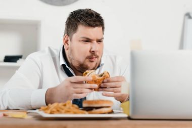 overweight businessman working while eating hamburger and french fries in office clipart