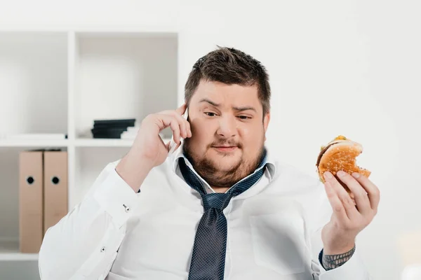 Übergewichtiger Geschäftsmann Spricht Auf Smartphone Während Büro Hamburger Isst — Stockfoto