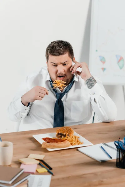 Übergewichtiger Geschäftsmann Telefoniert Arbeitsplatz Mit Dem Smartphone Und Isst Junk — Stockfoto