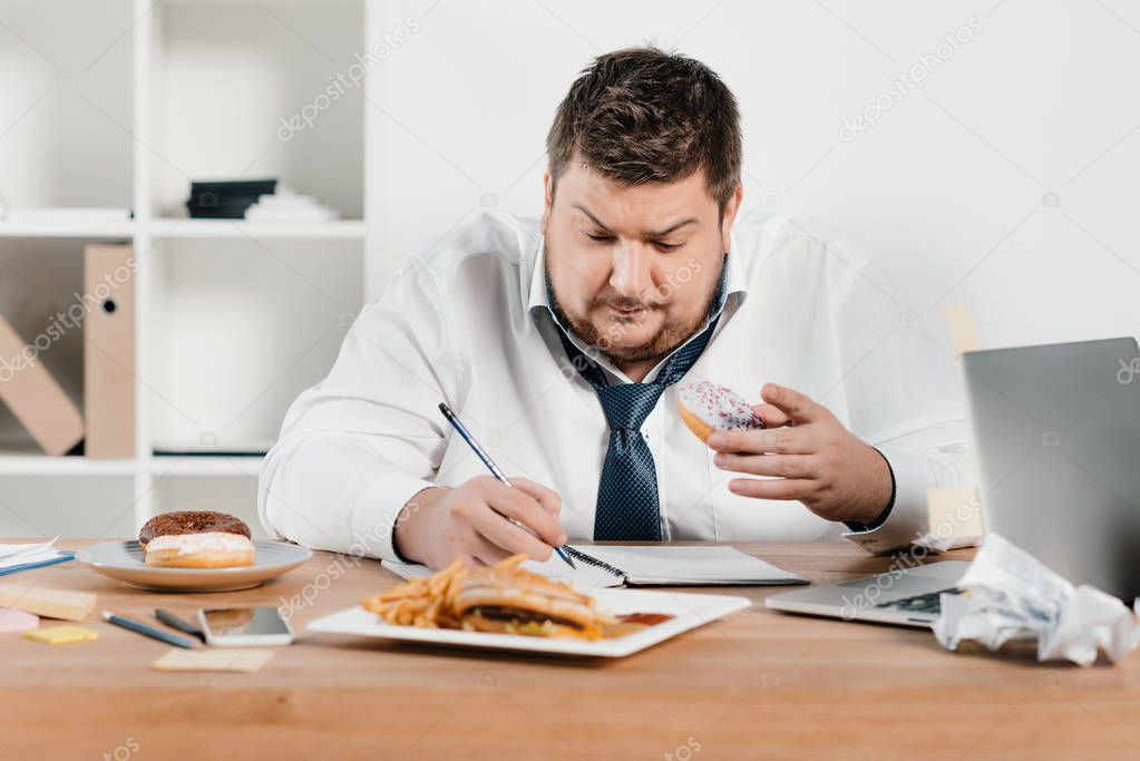 overweight businessman eating donuts, hamburger and french fries while wokring in office
