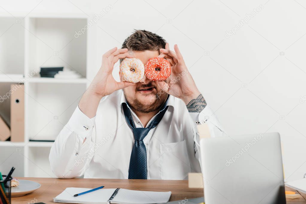 overweight businessman holding donuts in front of face in office