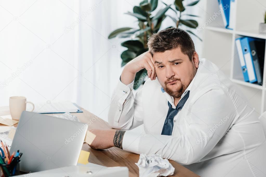 fat businessman sitting at workplace with crumpled papers and laptop