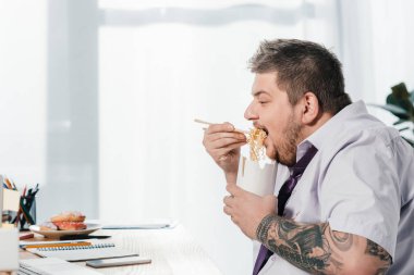 overweight businessman eating noodles at workplace in office clipart