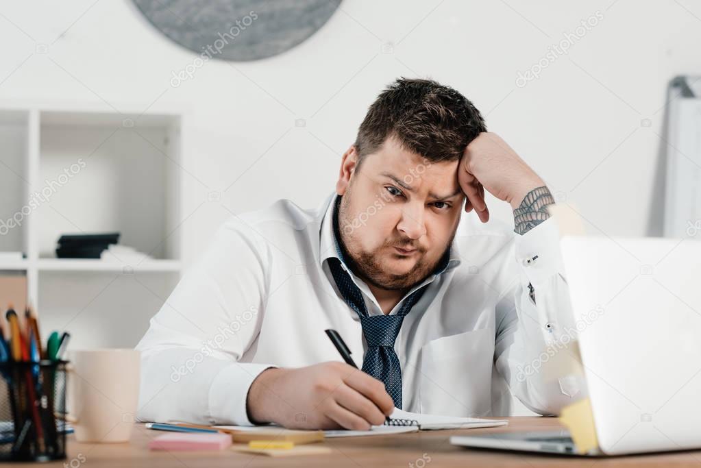 upset overweight businessman working with notepad and laptop in office