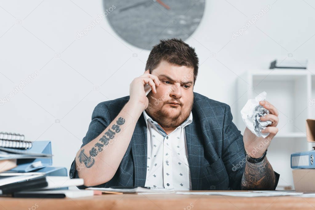 overweight businessman in suit talking on smartphone and working with documents in office