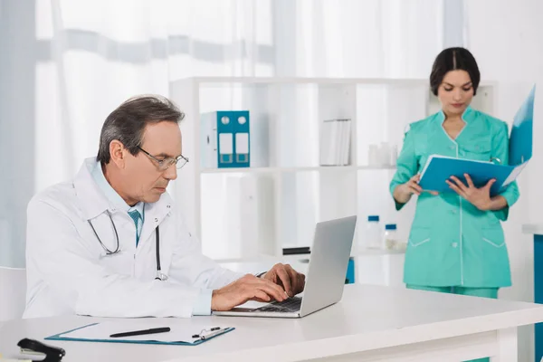 Pielęgniarka Szukając Czegoś Folderze Lekarz Pomocą Laptopa — Zdjęcie stockowe