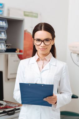 Kameraya bakarak elinde Not Defteri ile beyaz paltolu gülümseyen optometrist portresi