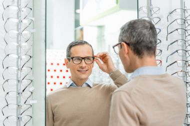 smiling caucasian man choosing pair of eyeglasses in optics clipart