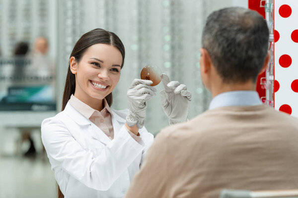 улыбающийся офтальмолог в белом халате показывает объектив пациенту в оптике
