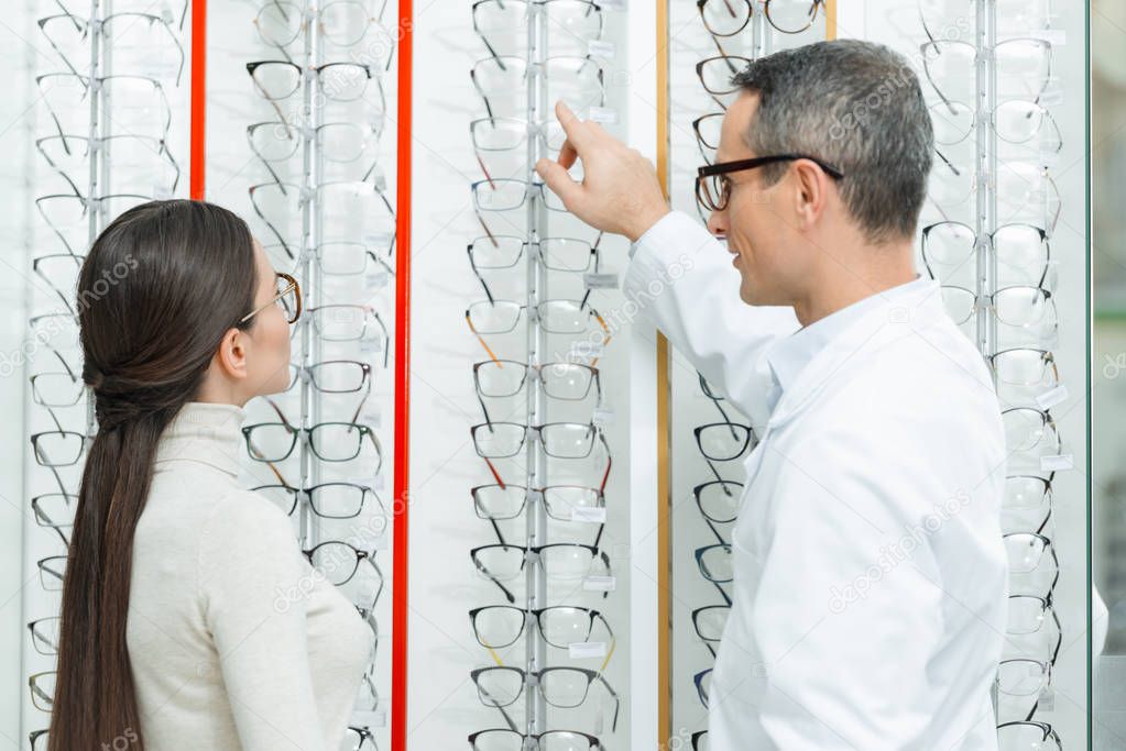 side view of oculist helping woman to choose pair of eyeglasses in optics