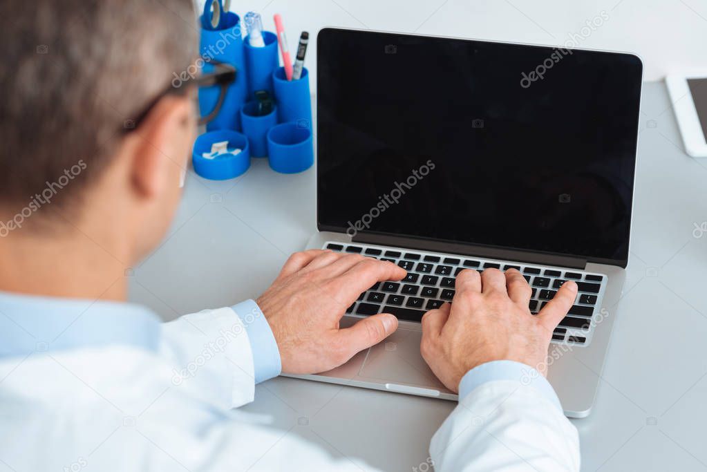 partial view of doctor working on laptop with blank screen at table in clinic