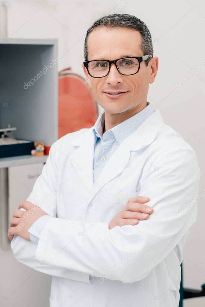 portrait of smiling doctor in eyeglasses with arms crossed looking at camera in clinic