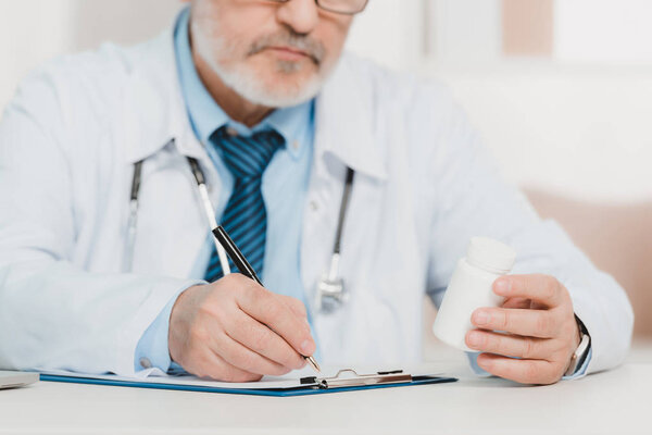 partial view of doctor with pills in hand making notes on notepad at workplace