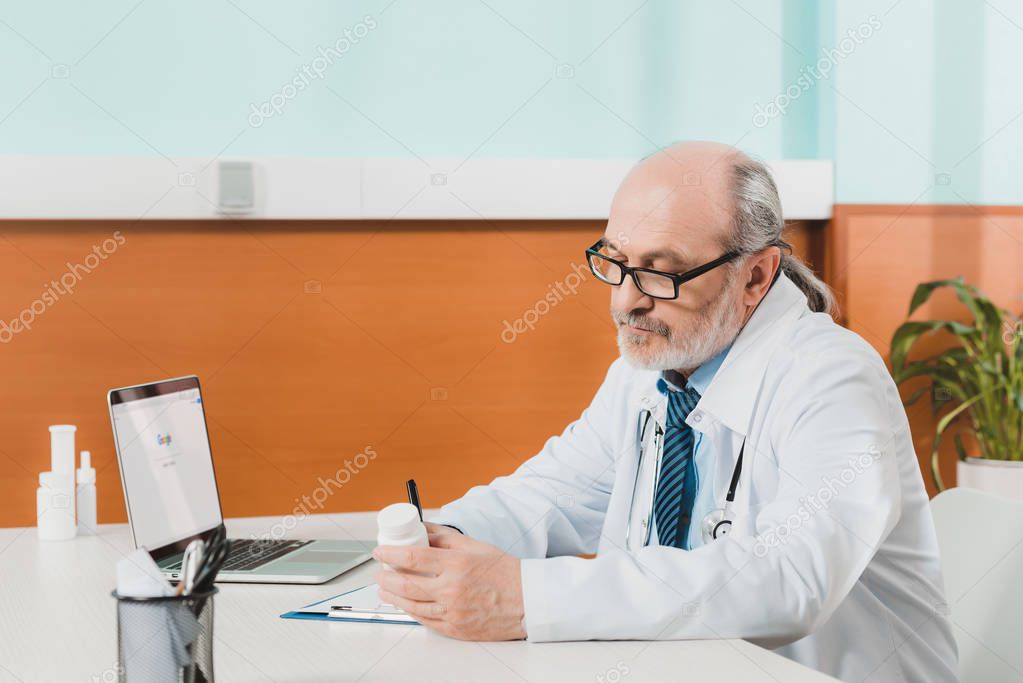 Side view of focused senior doctor making notes on notepad at workplace with laptop in clinic