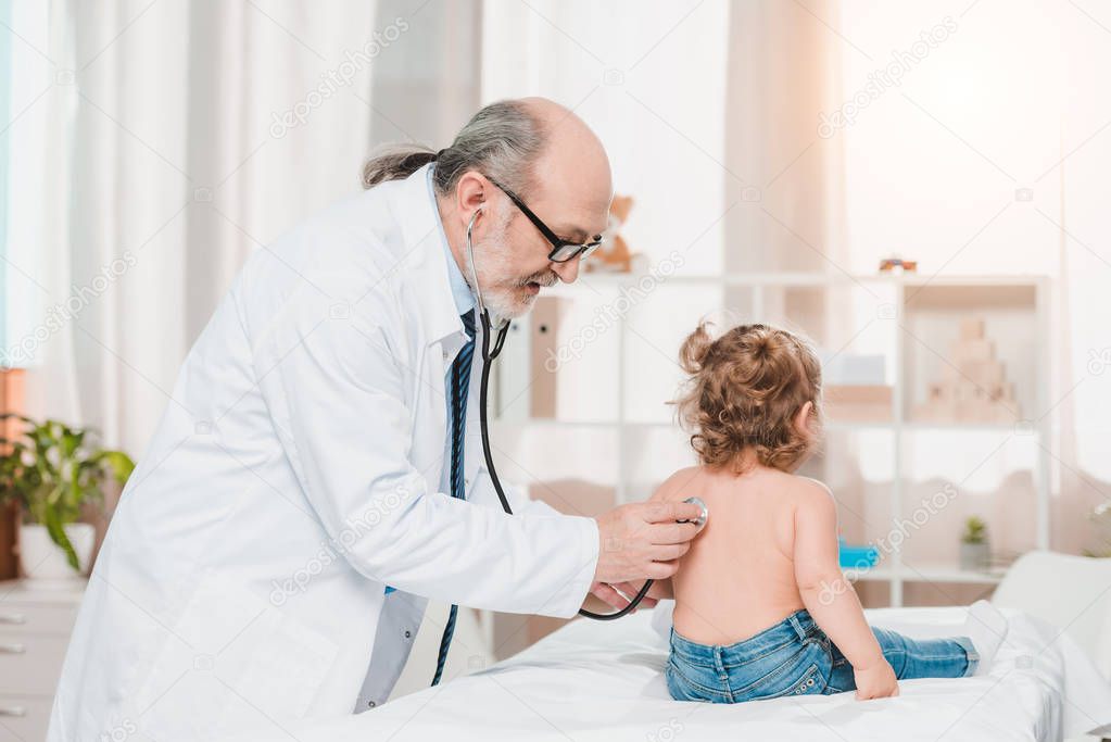 senior pediatrician in white coat examining little girl with stethoscope in clinic