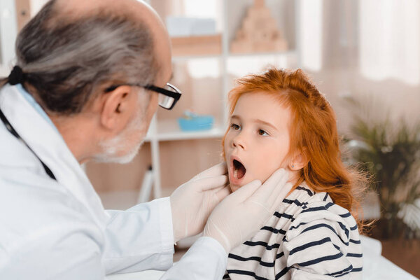 senior pediatrician in medical gloves examining little patients throat in clinic